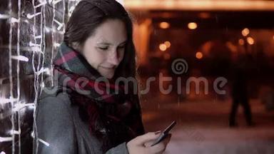 年轻迷人的女人在圣诞夜的大雪中使用智能手机站在灯光墙附近，